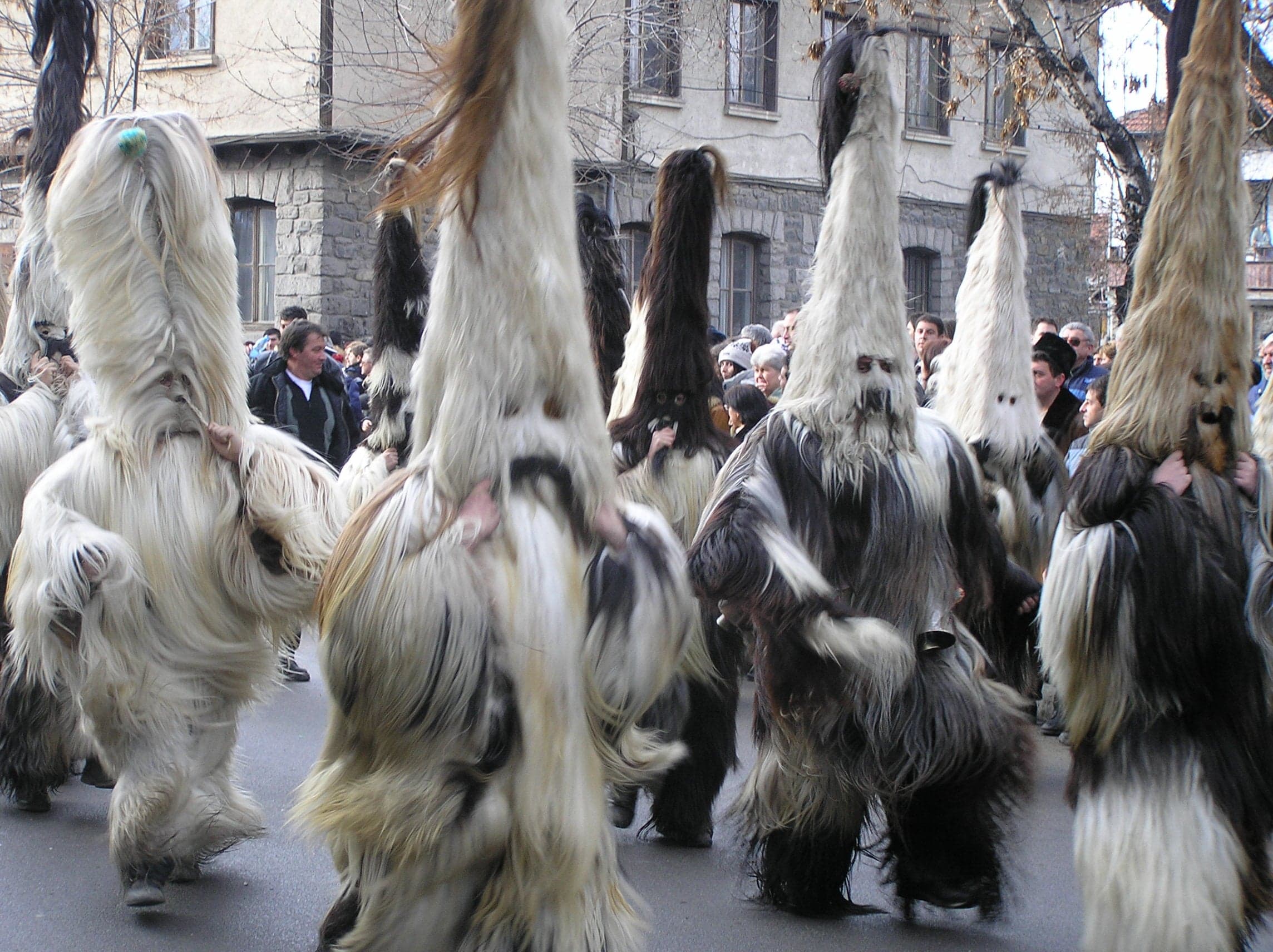 Kukeri performers in huge colourful fur costumes and carved wooden masks, with large bells around their waists, parading through a town square