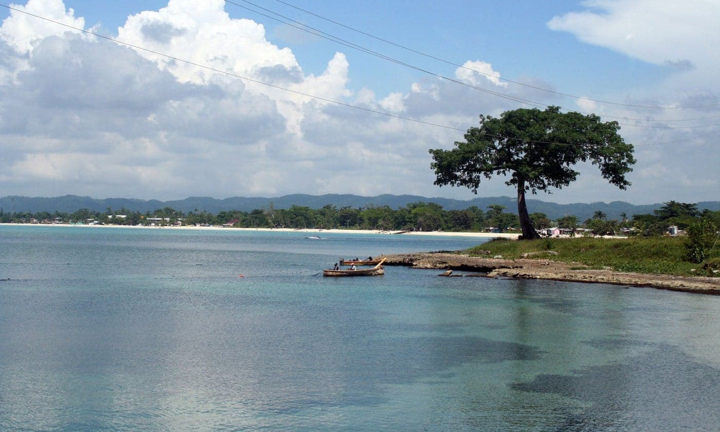 A wide, palm-fringed white sand beach stretching into the distance under a turquoise sky