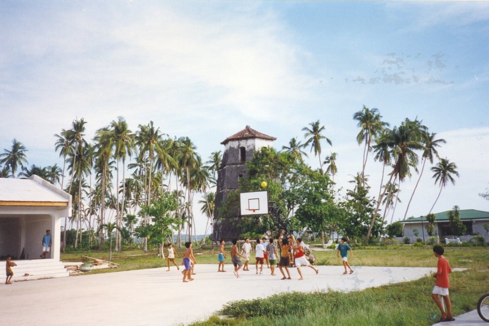 A group of Filipino children playing basketball on an outdoor court with palm trees and an old stone tower in the background