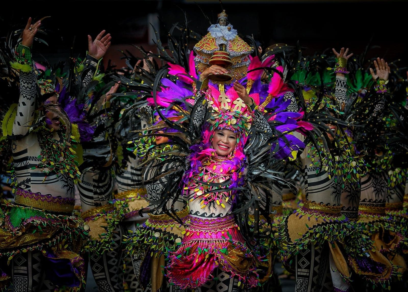 A Sinulog Festival dancer in a brilliant costume of pink, purple and green feathers, smiling with arms raised